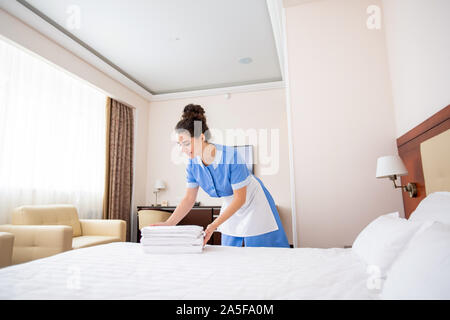 Pretty young professional chamber maid in uniform making stack of fresh white towels while bending over large clean bed Stock Photo