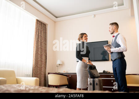 Young elegant porter with touchpad talking to pretty businesswoman with baggage while both standing in hotel room Stock Photo