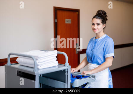 Pretty young brunette chamber maid pulling cart with clean towels while moving along corridor in hotel Stock Photo