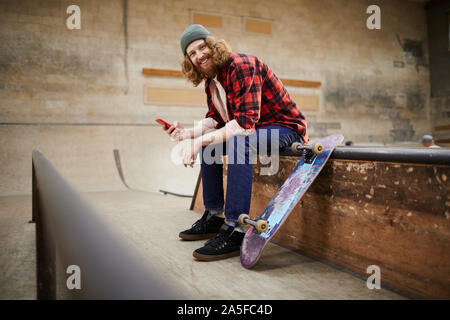 Full length portrait of contemporary bearded man looking at camera and smiling while using smartphone siting on ramp in skating park, copy space Stock Photo