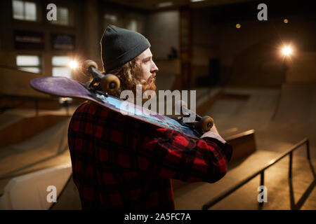 Back view portrait of contemporary bearded skater looking away pensively while posing in urban skating park lit by dim lights, copy space Stock Photo