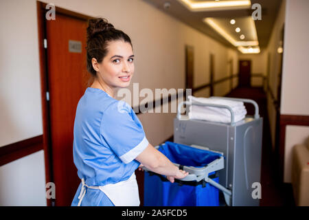 Young brunette chamber maid in uniform looking at you while pushing cart along corridor in hotel in the morning Stock Photo