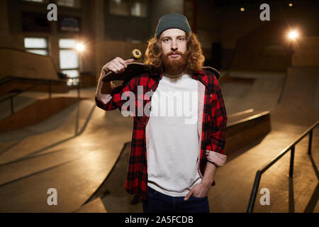 Waist up portrait of contemporary bearded man looking at camera while posing with skateboard in skating park, copy space Stock Photo