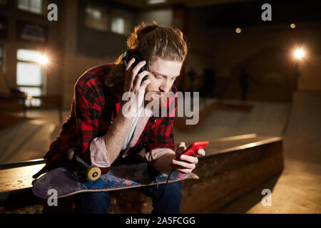 Portrait of contemporary bearded man listening to music via smartphone while sitting on ramp in dim skating park, copy space Stock Photo