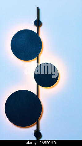 Stylish vertical metal wall lamp in the form of three celestial bodies in an eclipse against a blue wall makes soft warm lighting. Stock Photo