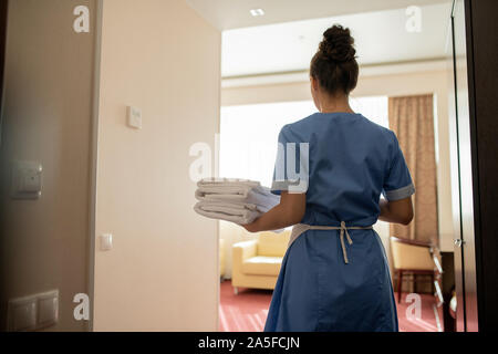 Back view of young brunette chamber maid in uniform carrying stack of clean white towels for guests while entering one of rooms Stock Photo