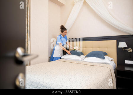 Young chamber maid in uniform putting pink flowers on one of pillows while making bed in the morning in hotel room Stock Photo