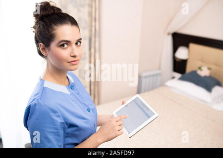 Pretty young chamber maid in uniform holding touchpad while working with electronic documents inside hotel room Stock Photo