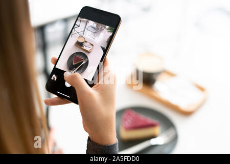 Cup of aromatic cappuccino and tiramisu cake on touchscreen of smartphone held by young woman relaxing in cafe Stock Photo
