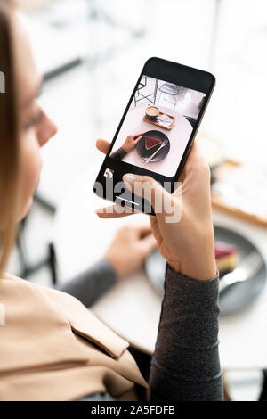 Female millennial with smartphone taking photo of fresh appetizing cheesecake and cup of cappuccino in cafe Stock Photo
