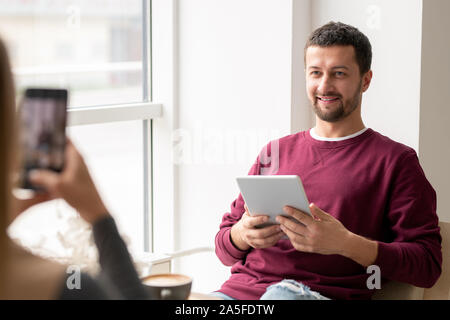 Smiling young man in casualwear looking at girlfriend taking his photo on smartphone while both relaxing in cafe Stock Photo