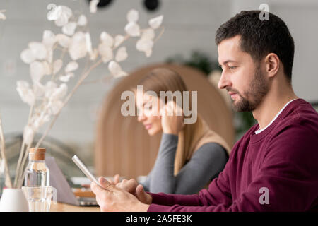 Serious man in casualwear scrolling in touchpad while sitting in cafe with young woman on background Stock Photo