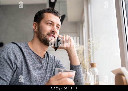 Happy young bearded man in pullover communicating to someone on mobile phone while relaxing in cafe Stock Photo