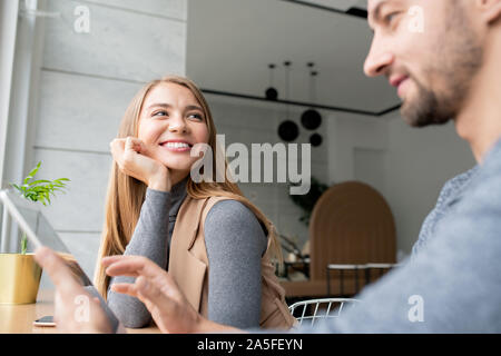 Young cheerful businesswoman sitting by her colleague using touchpad while both working over business project in cafe Stock Photo