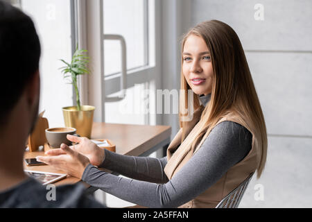 Young elegant blonde woman explaining something to her boyfriend or colleague while both sitting in cafe Stock Photo