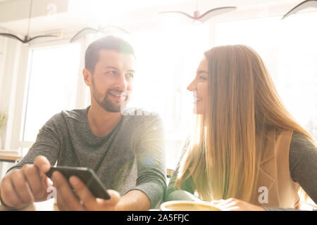 Happy young affectionate couple discussing funny stuff in cafe while scrolling or watching something in smartphone Stock Photo