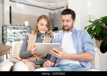 Casual young couple sitting on comfortable couch and scrolling in touchpad while relaxing in cozy cafe Stock Photo