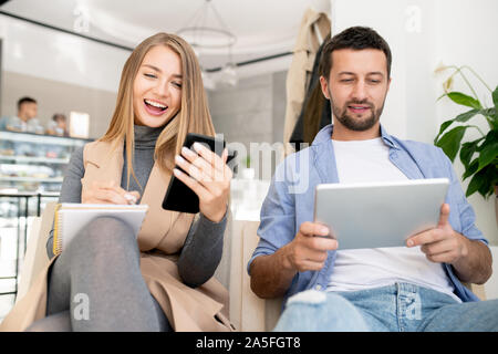 Happy young student looking at smartphone screen while making notes in notepad with her boyfriend using touchpad near by Stock Photo