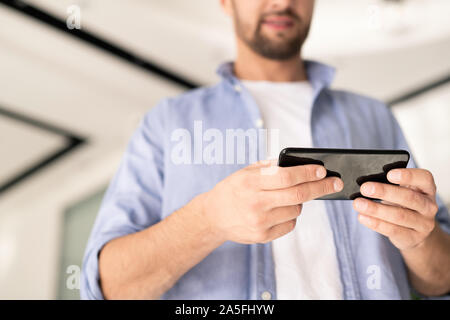 Young businessman in casualwear holding smartphone while watching something or communicating through video-chat Stock Photo
