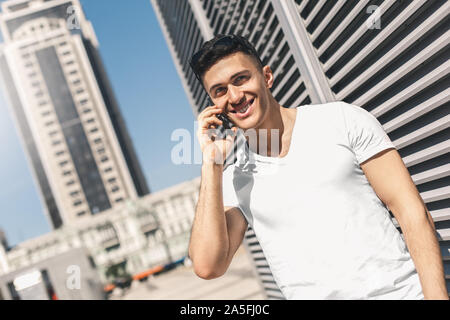 Outdoors Leisure. Stylish guy with sunglasses on head standing on the street talking on smartphone smiling happy Stock Photo