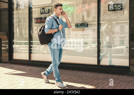 Outdoors Leisure. Stylish guy with backpack walking on the street talking on phone looking forward pensive side view Stock Photo