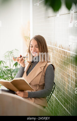 Happy young student reading notes in copybook and recording voice message for her friend while sitting in cafe Stock Photo
