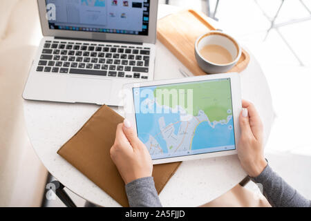 Young woman holding touchpad with map on its display while sitting by table, having cappuccino and using gadgets Stock Photo