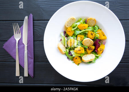 Delicious healthy salad with greens, pumpkin and sumac spices in big white plate on black table background. Violet napkin and cutlery Stock Photo