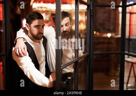 Portrait of bearded man carrying drunk businessman through door while leaving bar late night after work, copy space Stock Photo