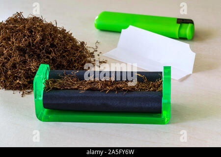 Green cigarette rolling machine, cigarette paper, green lighter and pile of tobacco on a white table. Making cigarettes with pipe tobacco at home. Stock Photo