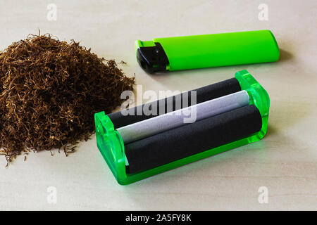 Green cigarette rolling machine, green lighter and pile of tobacco on a white table. Making cigarettes with pipe tobacco at home. Front view. Stock Photo