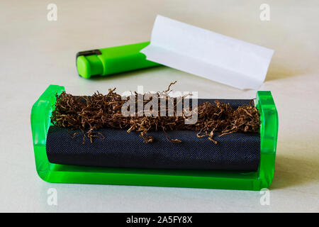 Green cigarette rolling machine with tobacco, cigarette paper and green lighter on a white table. Making cigarettes with pipe tobacco at home. Stock Photo