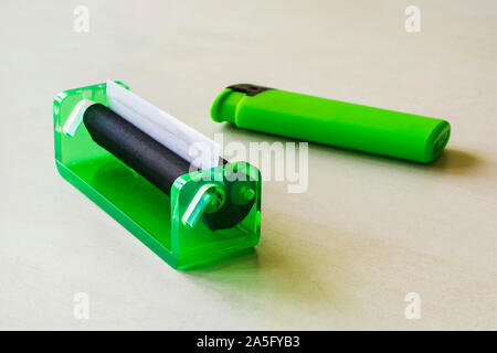 Green rolling machine with alwost done cigarette and green lighter on a white table. Making cigarettes with pipe tobacco at home. Front view. Stock Photo
