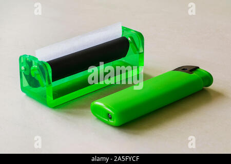 Green lighter and green rolling machine with alwost done cigarette on a white table. Making cigarettes with pipe tobacco at home. Front view. Stock Photo