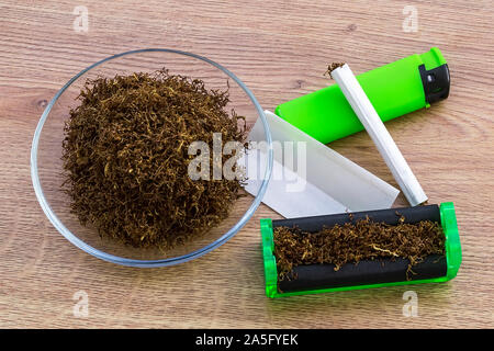 Pile of tobacco in glass saucer, green cigarette rolling machine, paper, one cigarette and green lighter on a wooden table. Making cigarettes. Stock Photo