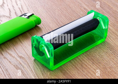 Green lighter and green rolling machine with alwost done cigarette on a wooden table. Making cigarettes with pipe tobacco at home. Front view. Stock Photo