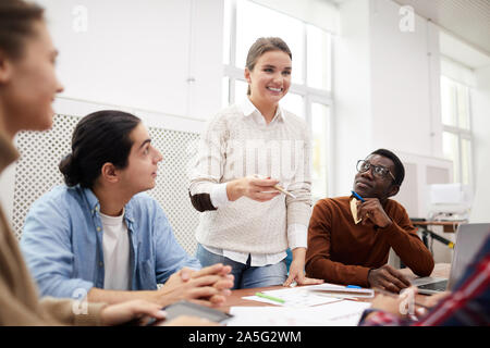 Multi-ethnic group of students working together on team project while studying in college, focus on smiling girl heading meeting Stock Photo