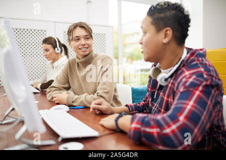 College students sitting in row and chatting while studying in computer class, focus on smiling young woman talking to friend, copy space Stock Photo