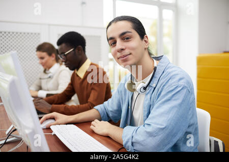 Portrait of young Latin-American man smiling at camera while using computer in college library, copy space Stock Photo