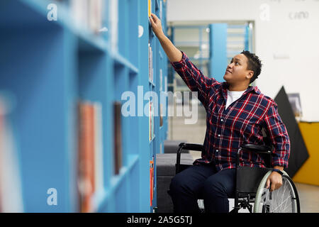 Portrait of disabled student in wheelchair choosing books while studying in college library, copy space Stock Photo