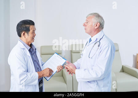 Senior multi-ethnic general practitioners in white lab coats talking in hall of medical clinic Stock Photo