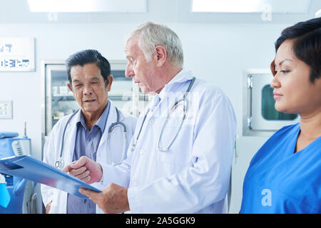 Senior general practitioner discussing document on clipboard with Vietnamese coworker Stock Photo