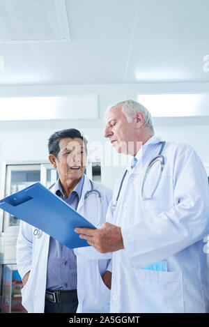 Positive Vietnamese general practitioner looking at his coworker reading medical history of patient on tablet computer Stock Photo