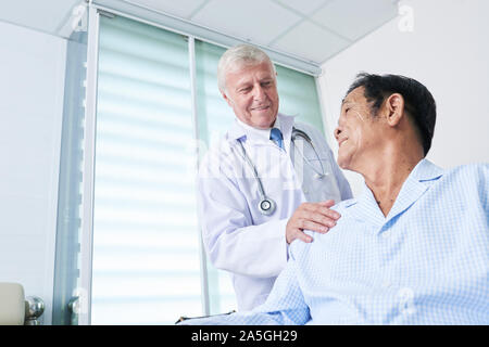 Smiling general practitioner patting shoulder of senior Vietnamese man to support him during recovery therapy Stock Photo