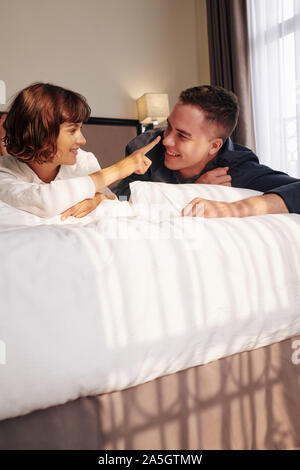 Pretty young smiling woman lying in bed with her boyfriend and pointing at his nose Stock Photo