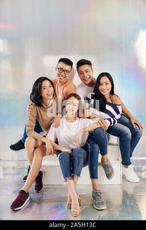 Cheerful young Asian people in casual clothes posing together on steps in studio with holographic background Stock Photo