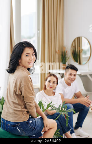 Pretty young Asian woman attending meeting with friends, sitting on sofa and turning back to camera Stock Photo