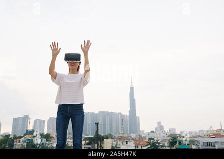 Happy young woman standing outdoors and raising hands to touch something when experiencing virtual reality Stock Photo