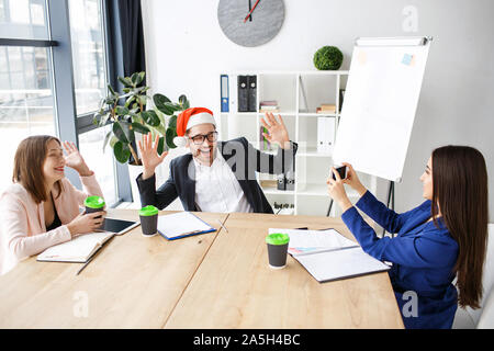 Workers in office. Celebrating new year or Christmas. People sit at table together. Brunette taking picture of man in red hat. Having fun at work Stock Photo