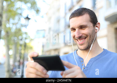 Happy adult man wearing earbuds watching videos on horizontal mobile phone in the street Stock Photo